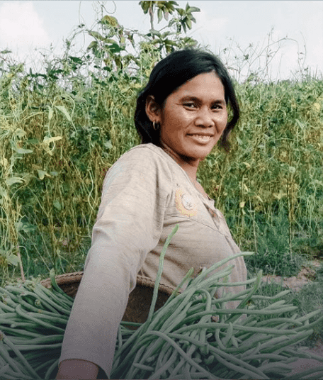 An Asian woman farmer in the middle of a field is looking straight at the camera and smiling. She is holding a basket full of green crops.