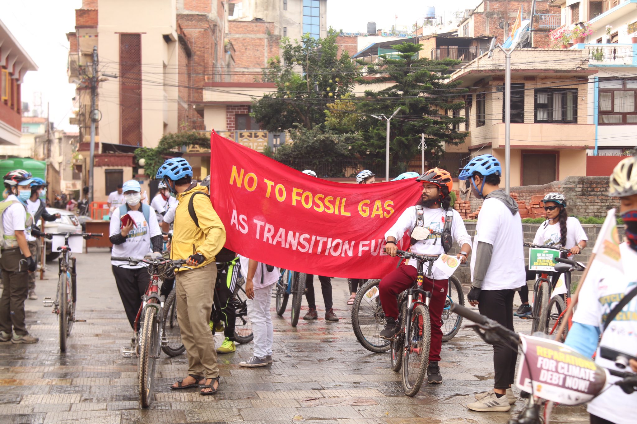 A bike rally where a bunch of cyclists and climate activists are all gathered together on a street in Kathmandu, Nepal. A big red banner with the words "No to fossil gas as transition fuel" in yellow font is written. Photo by Digo Bikas Institute. 