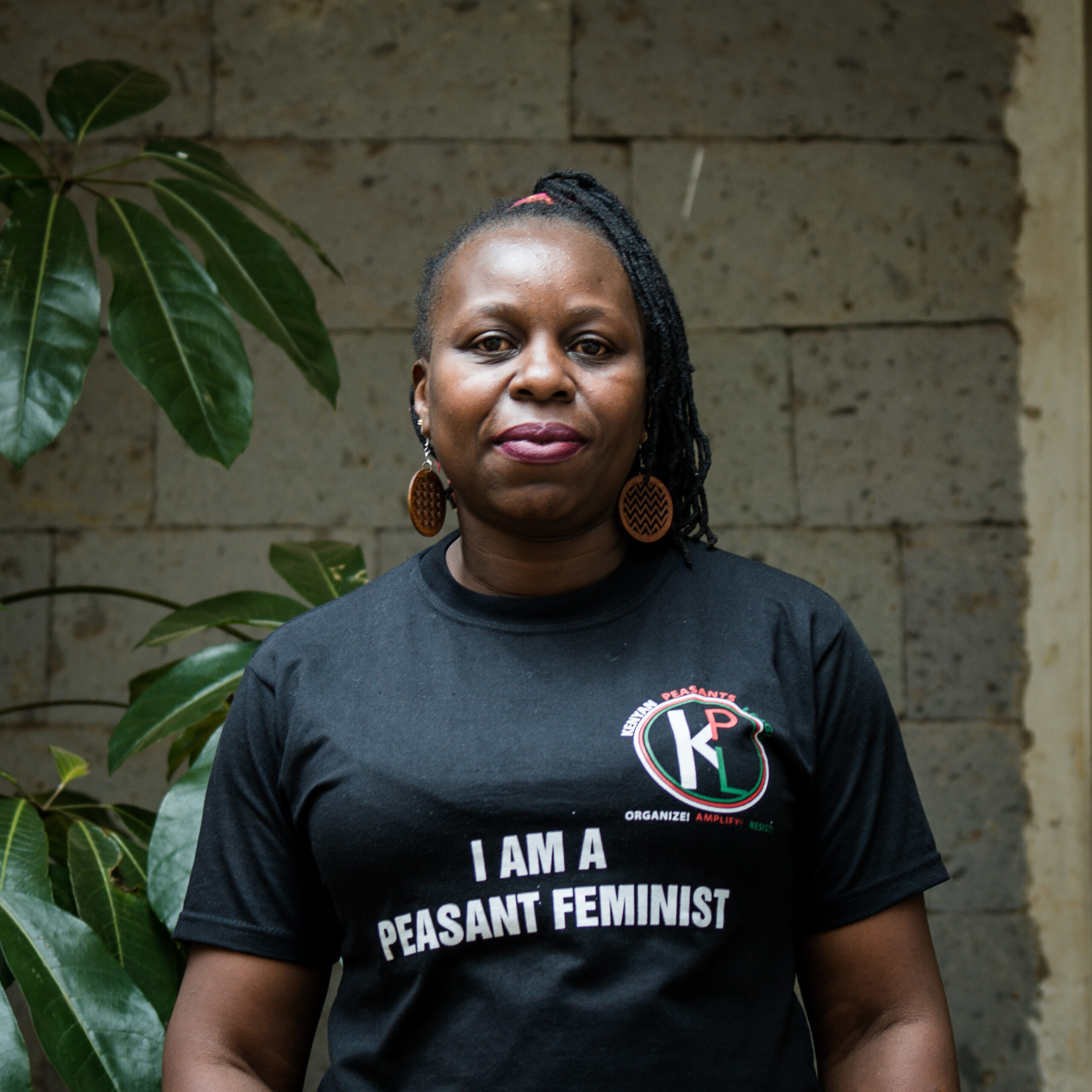 A close up shot of a Black woman. She is wearing a black t-shirt that says "I am a peasant feminist" with the KPL logo on the top right. She's staring at the camera with a slight smile. Few leaves are visible in the background. Pic credit: Ayşe Gürsöz